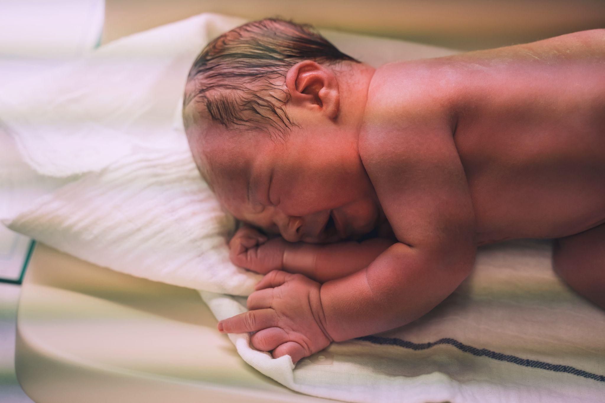 Crying newborn baby lying on their side on hospital blanket. Vernix is still on their back.