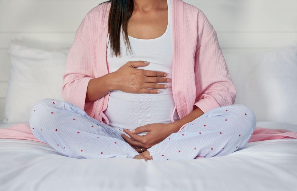 A pregnant woman in pyjamas relaxes cross-legged on her bed with her hands on her bump.