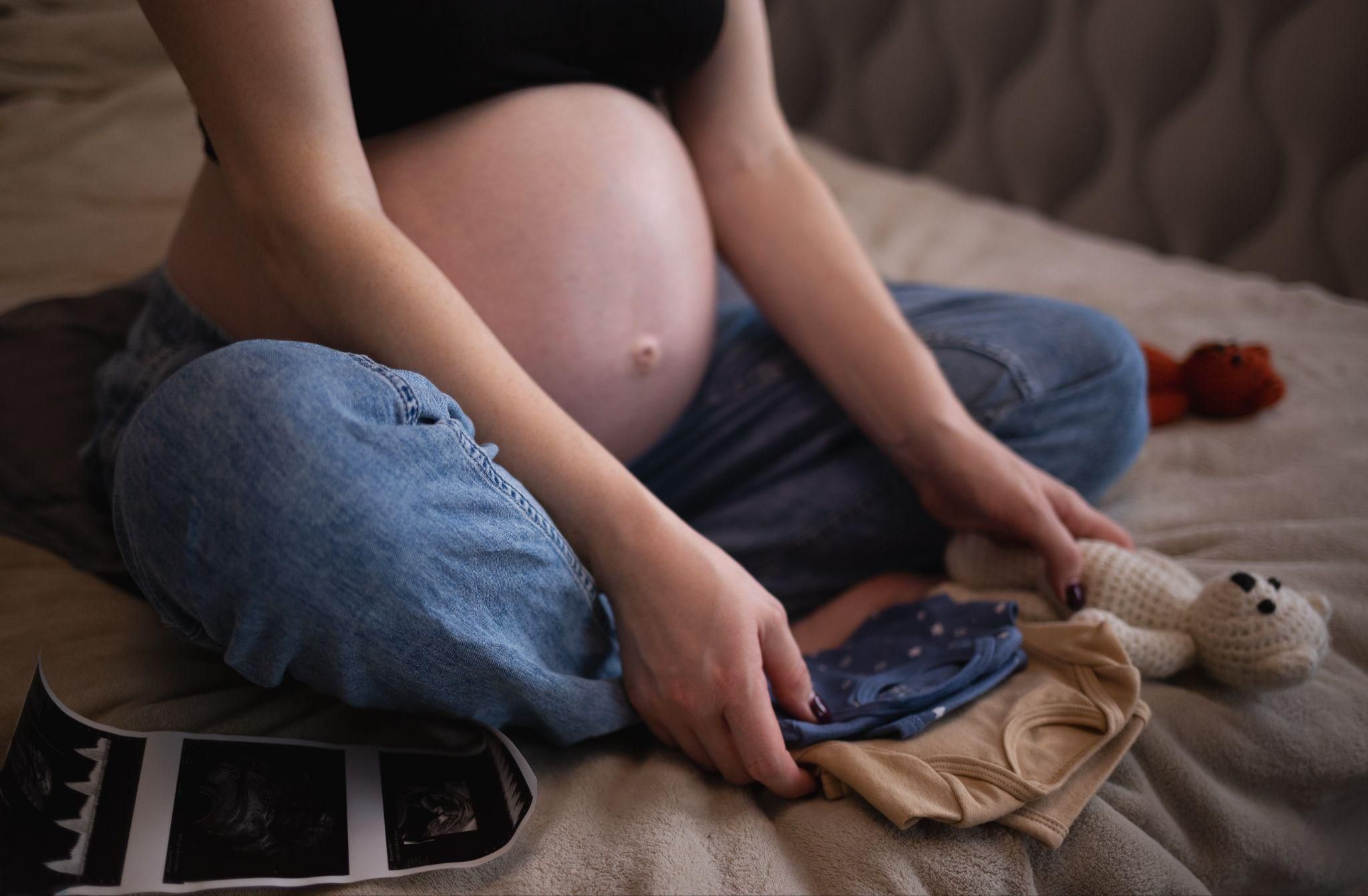 Pregnant lady sits cross-legged on bed with folded baby clothes, knitted teddy bears, and ultrasound photos.