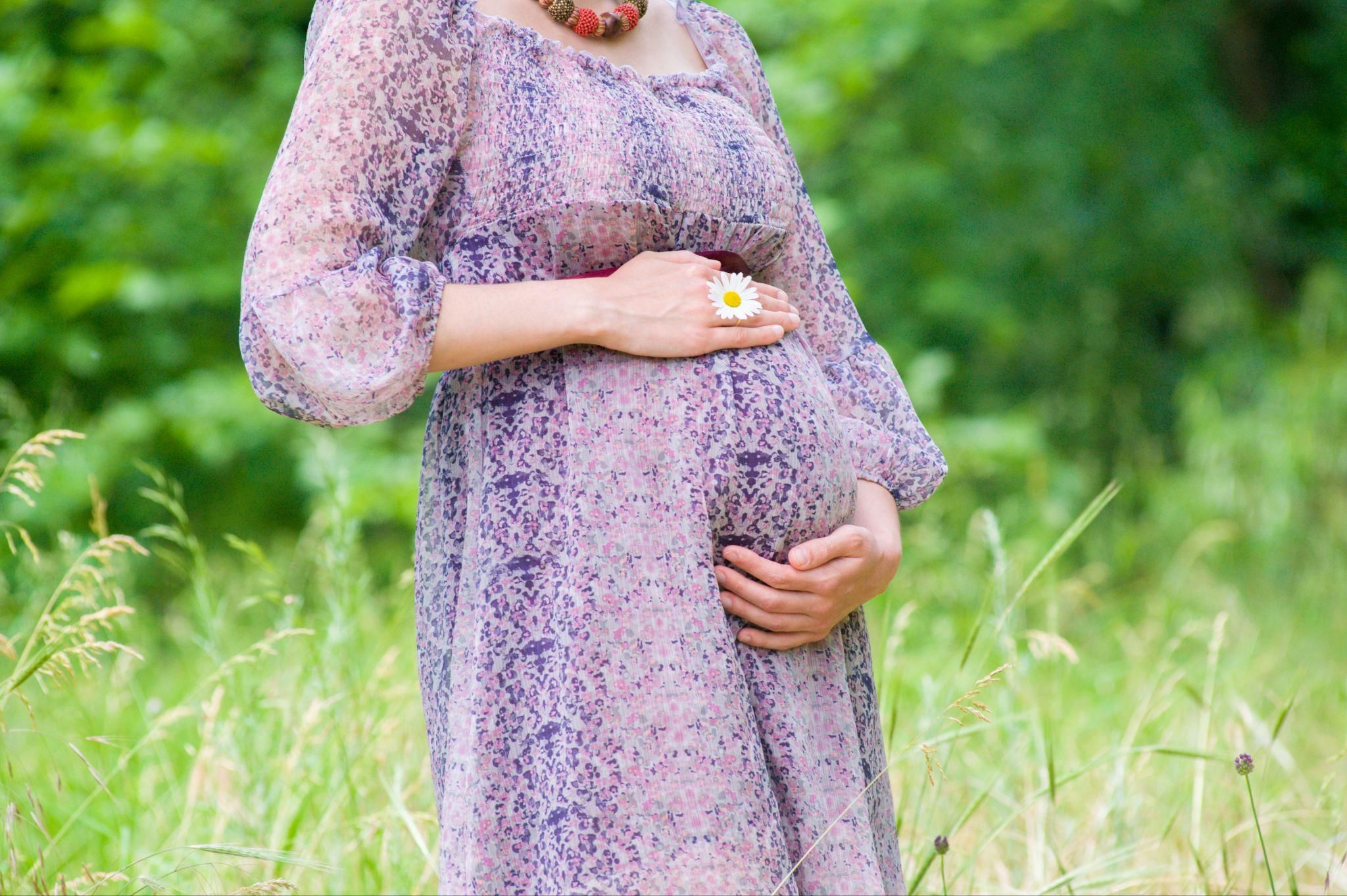 Pregnant lady with hands on baby bump stands in sunny field.