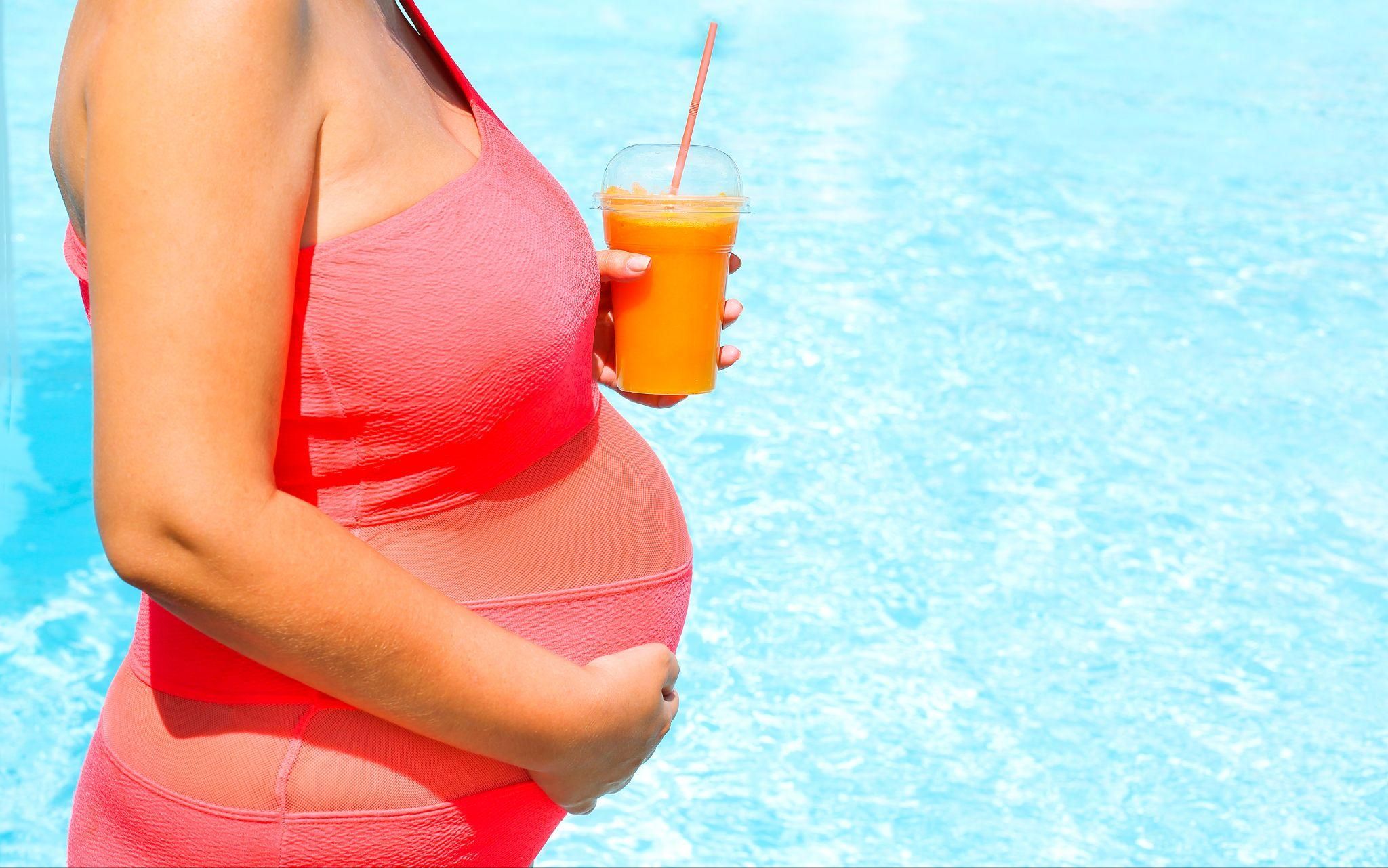 A pregnant woman drinks a fruit smoothie by a swimming pool.