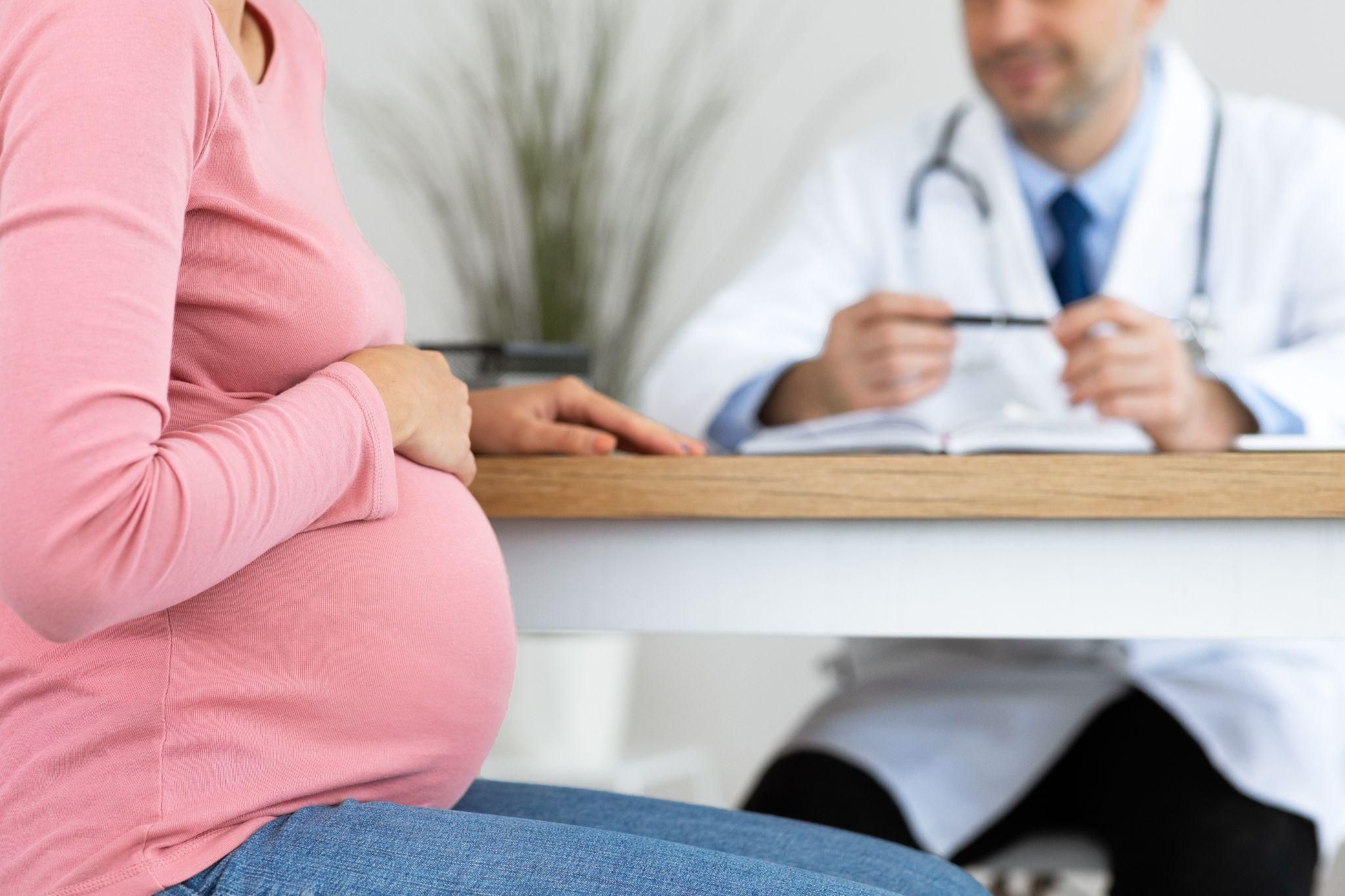 A pregnant woman sits speaking with a male obstetrician and gynaecologist at his desk during an antenatal clinic consultation.