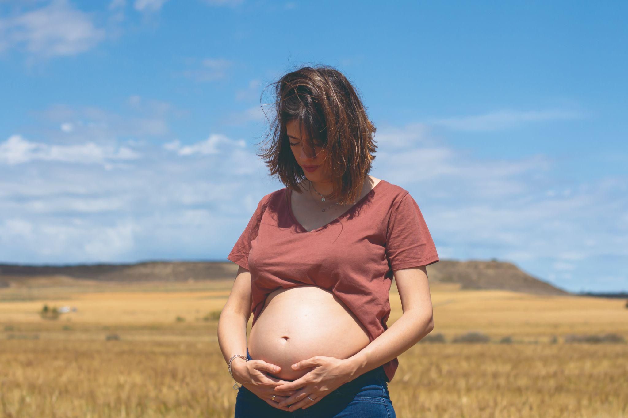 Pregnant woman stands outdoors in a bucolic environment with her hands supporting her bump.