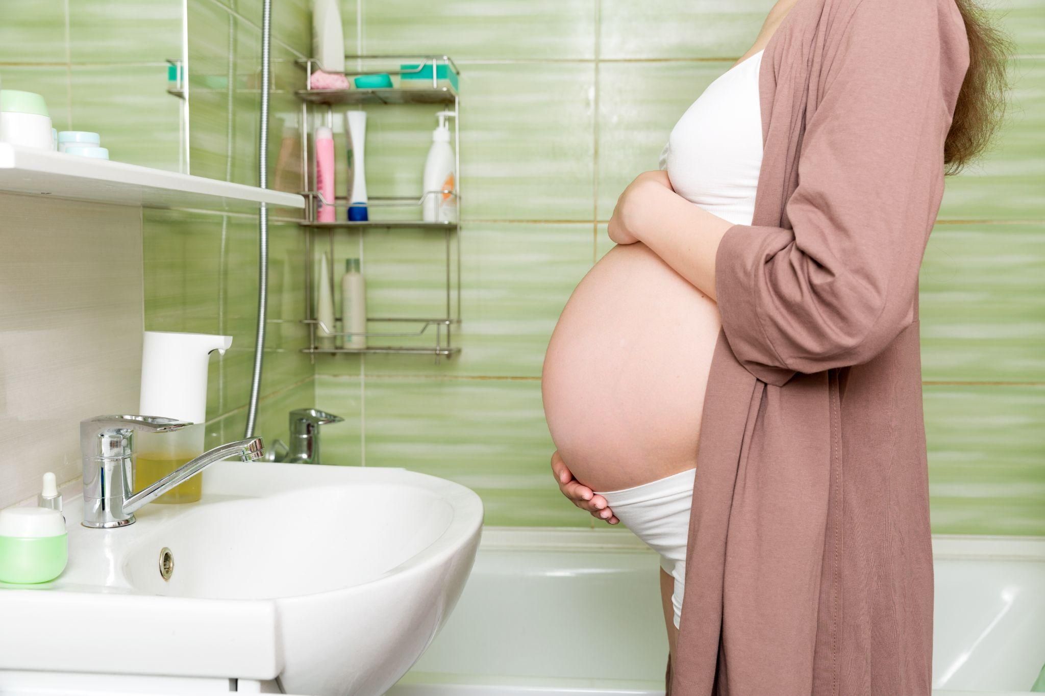 Pregnant woman in dressing gown at bathroom sink, holding her bump.