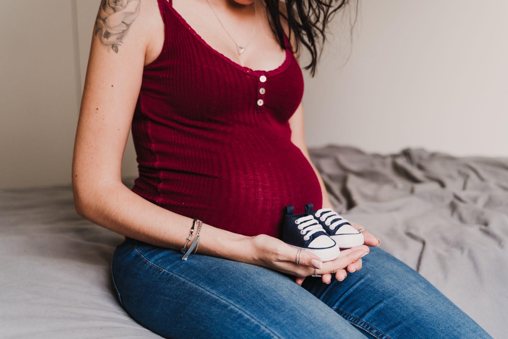 Pregnant woman rests comfortably on bed with baby shoes in hands.