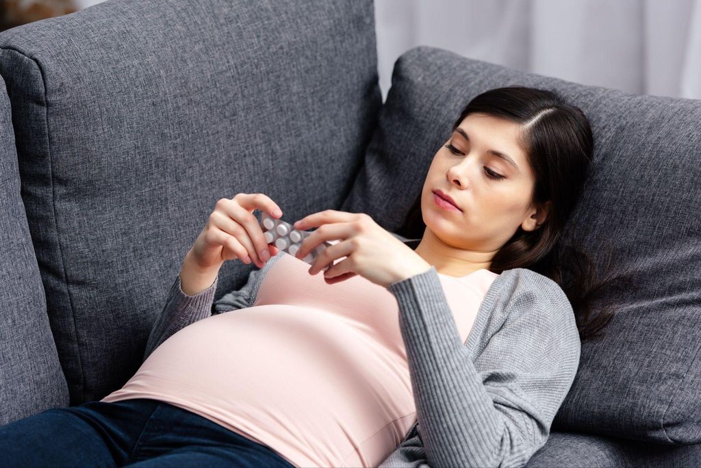 Pregnant woman examines sheet of pills while lying on the sofa.