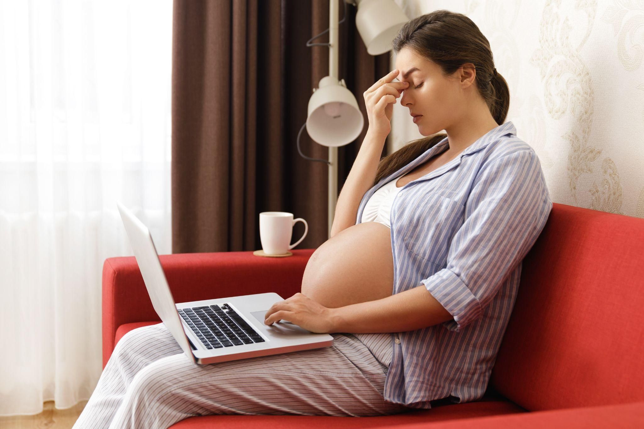 Pregnant woman pinches bridge of nose with worry while Googling pregnancy symptoms on laptop and sitting in pyjamas on couch in daytime. Possible case of antenatal anxiety.