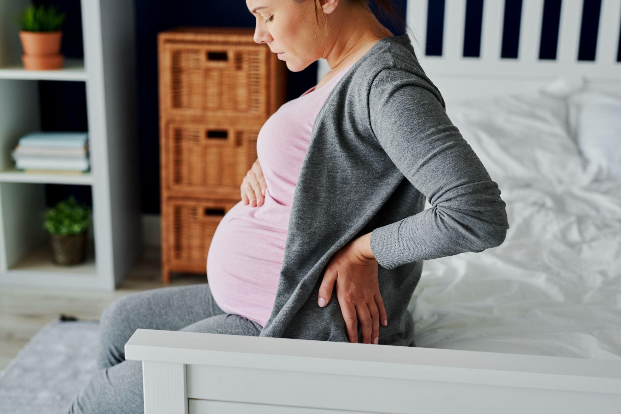 Pregnant woman with large bump sits on bed, wincing from antenatal back pain.