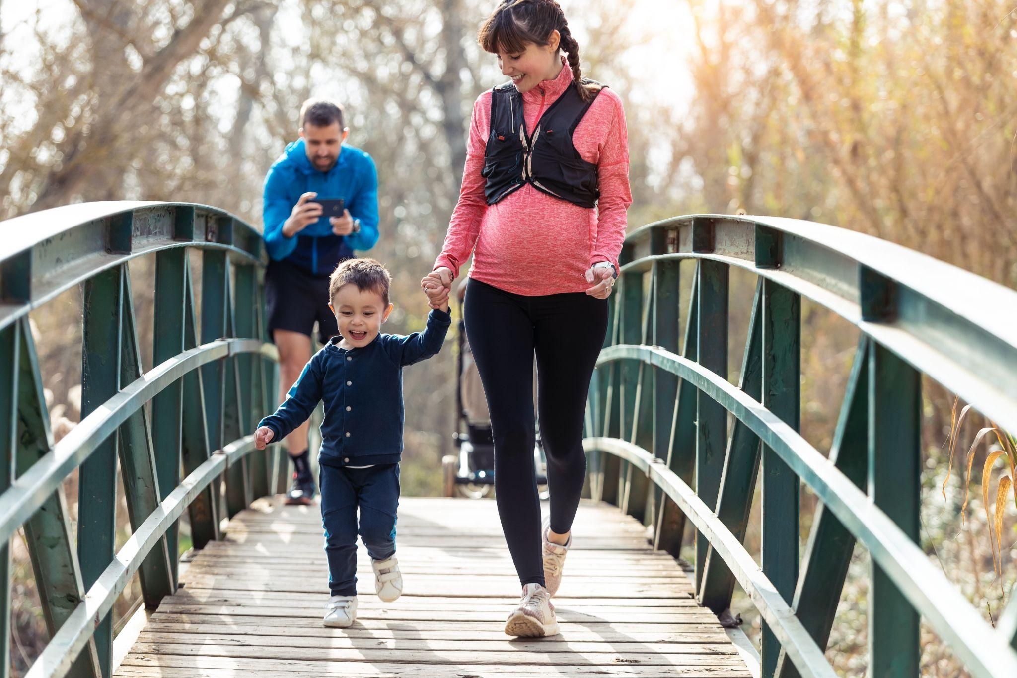 Pregnant woman in workout clothes and supportive footwear walks outdoors with young son and partner.