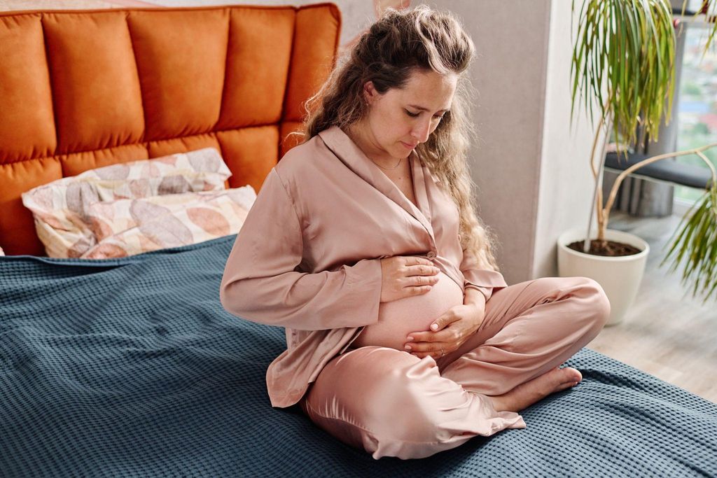 A pregnant woman sits cross legged on her bed in pink pyjamas, rubbing her bump.