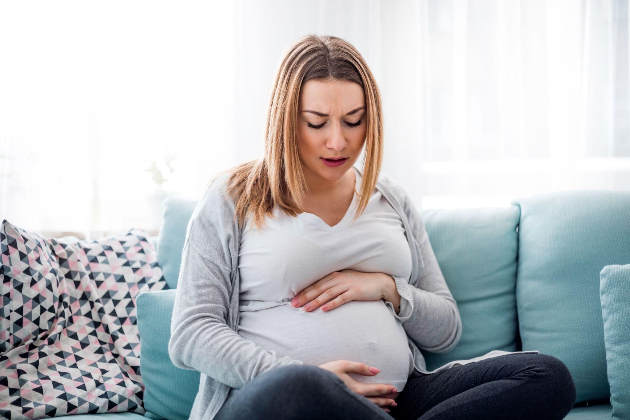 Stressed pregnant woman sits on couch with hands on belly. Possible case of antenatal depression or anxiety.