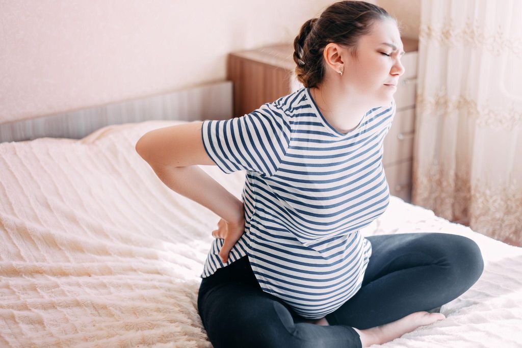 A pregnant woman with a large bump suffers lower back pain and sits cross-legged on bed.