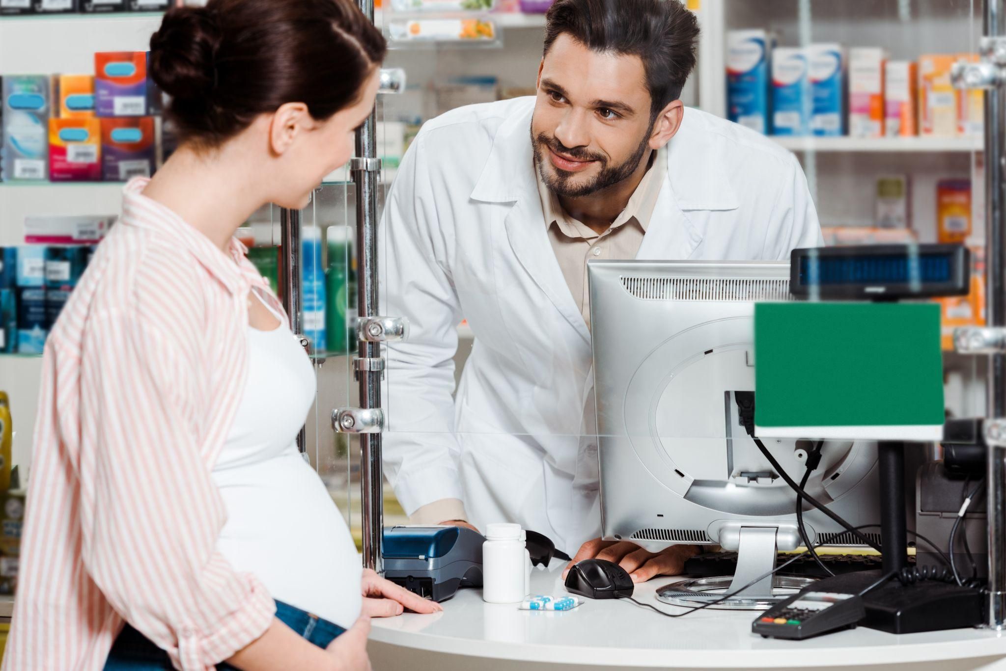 Male pharmacist chats to pregnant woman about medication in chemist at counter.