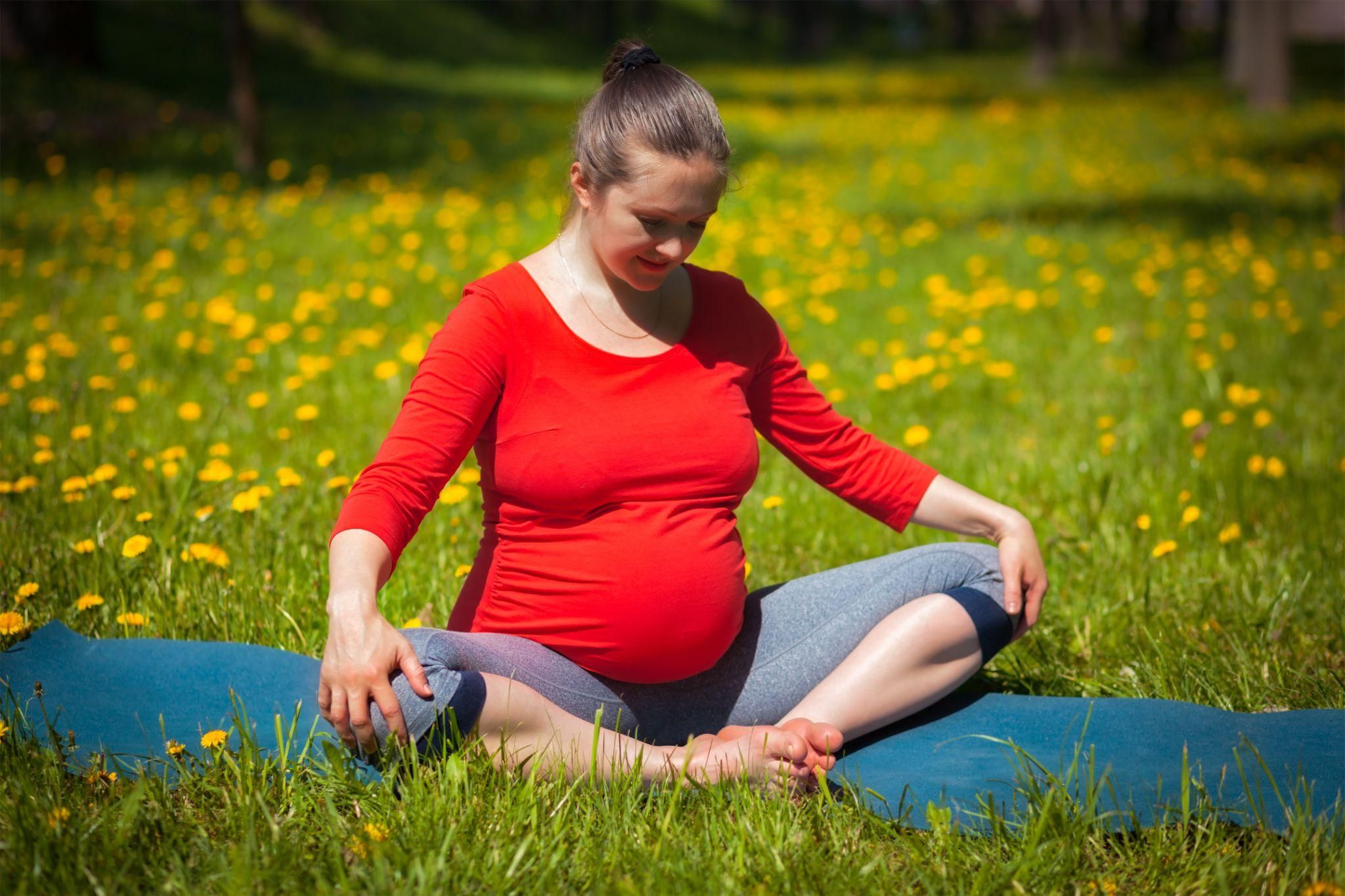 A pregnant woman performs a gentle yoga pose in her third trimester while sitting on a yoga mat outdoors.