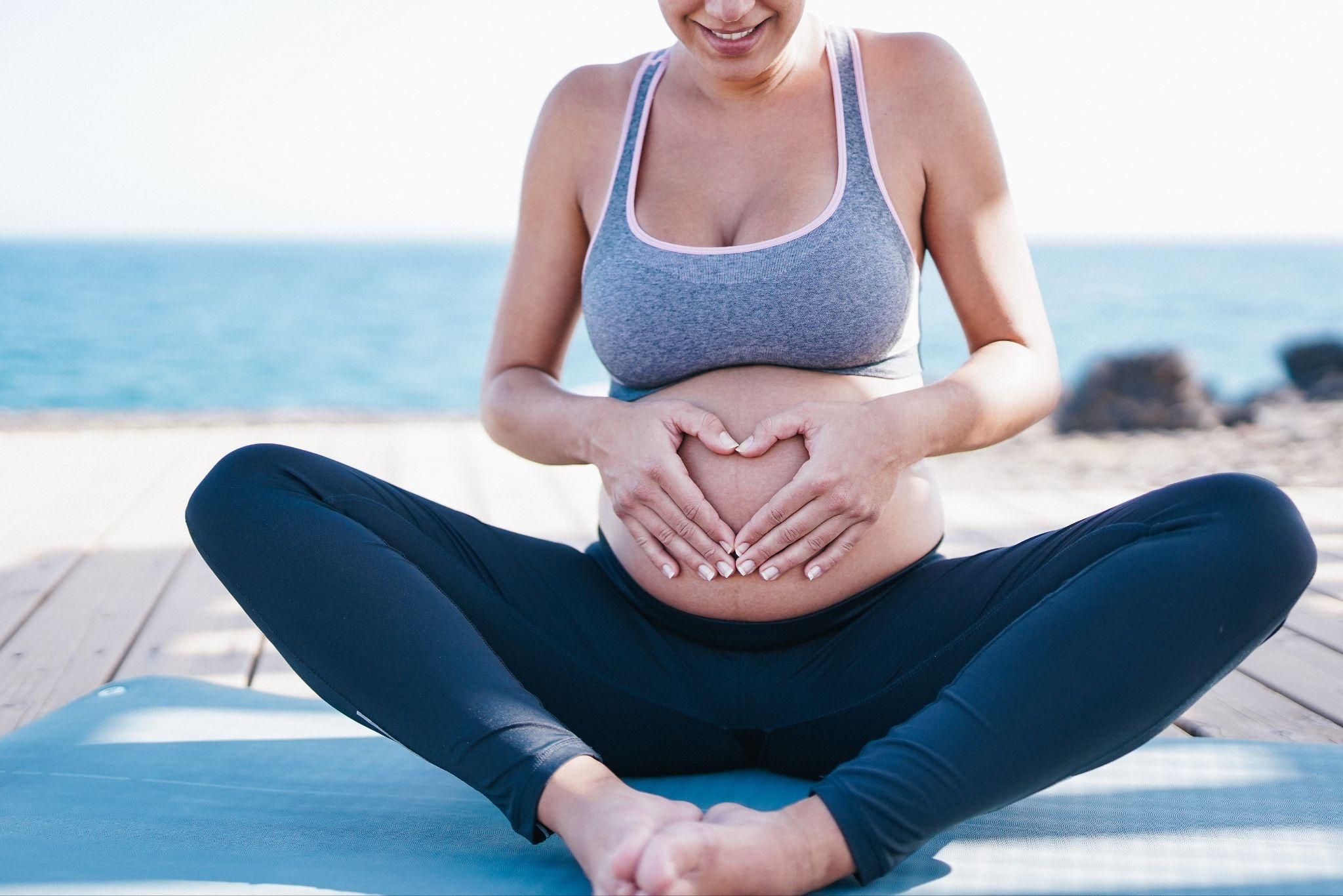 Pregnant woman sits on yoga mat outdoors by the sea while smiling and holding her bump.