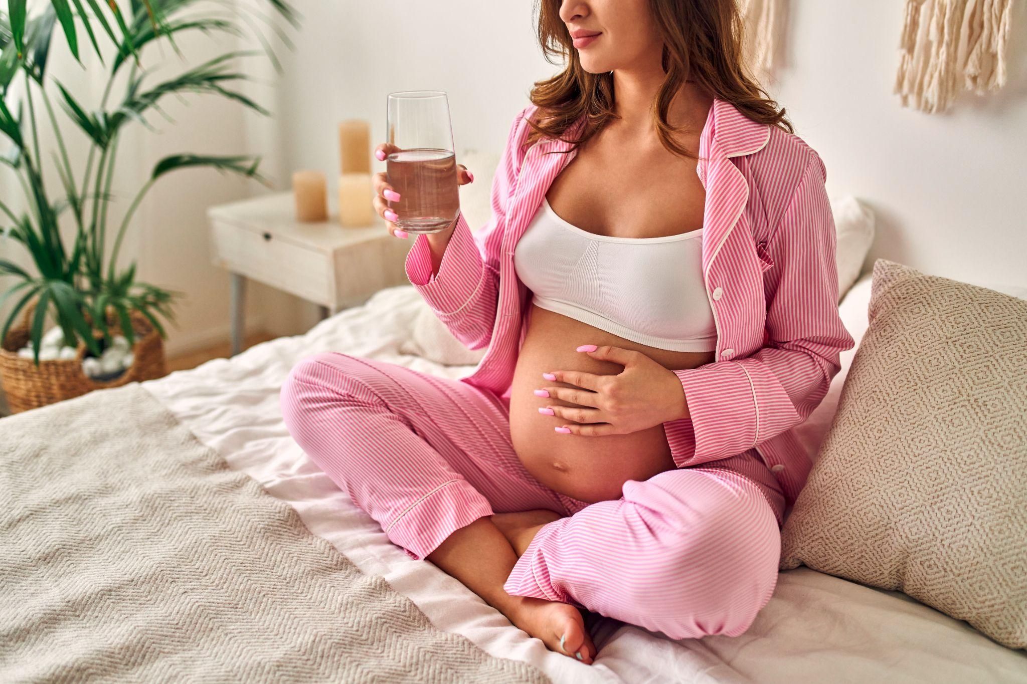 A pregnant woman sits in her pyjamas on her bed drinking a glass of water.