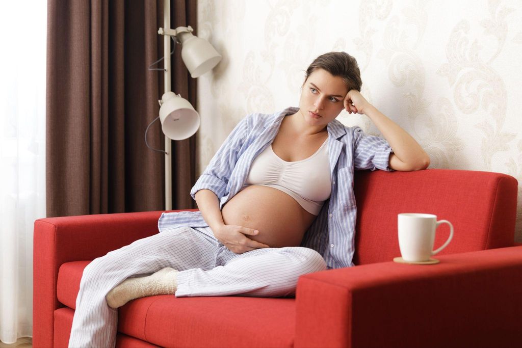 Sad pregnant woman who may have antenatal depression rubs belly while she sits on sofa in pyjamas.