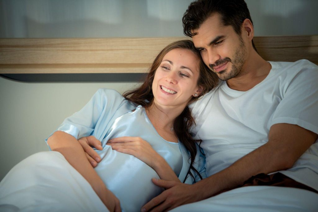 A pregnant couple smiling and embracing in bed, discussing antenatal sex and relationship changes.