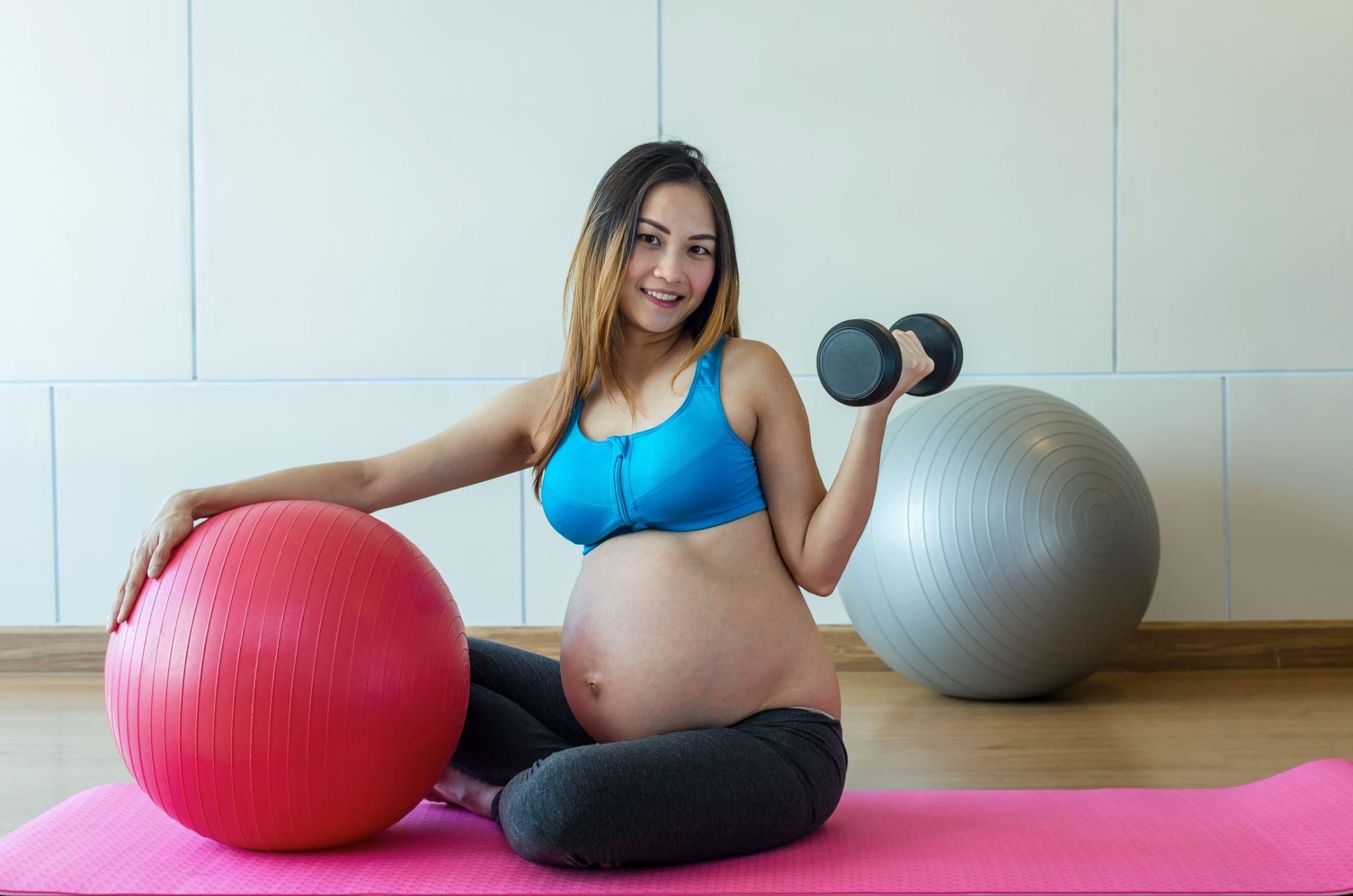 Pregnant woman lifts weights, holds a birthing ball, and sits on a yoga mat in the gym.