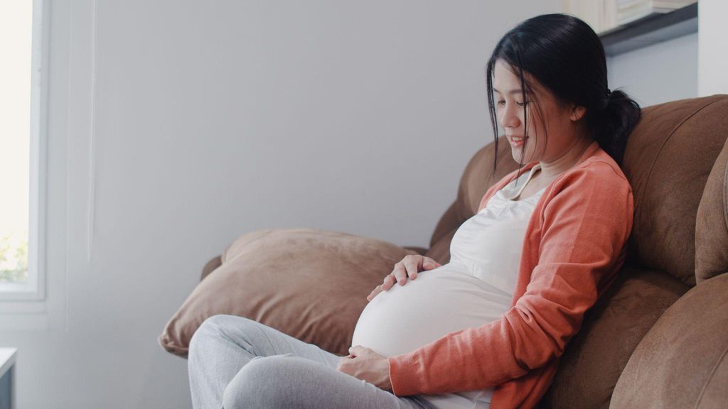 A mum-to-be sitting on a sofa, noting her baby’s antenatal movement patterns as she smiles.