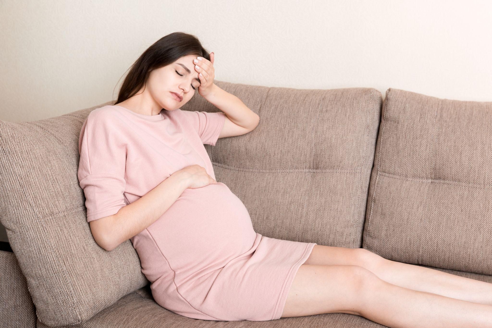 Pregnant woman sits on couch looking tired and stressed. Possible case of antenatal depression.