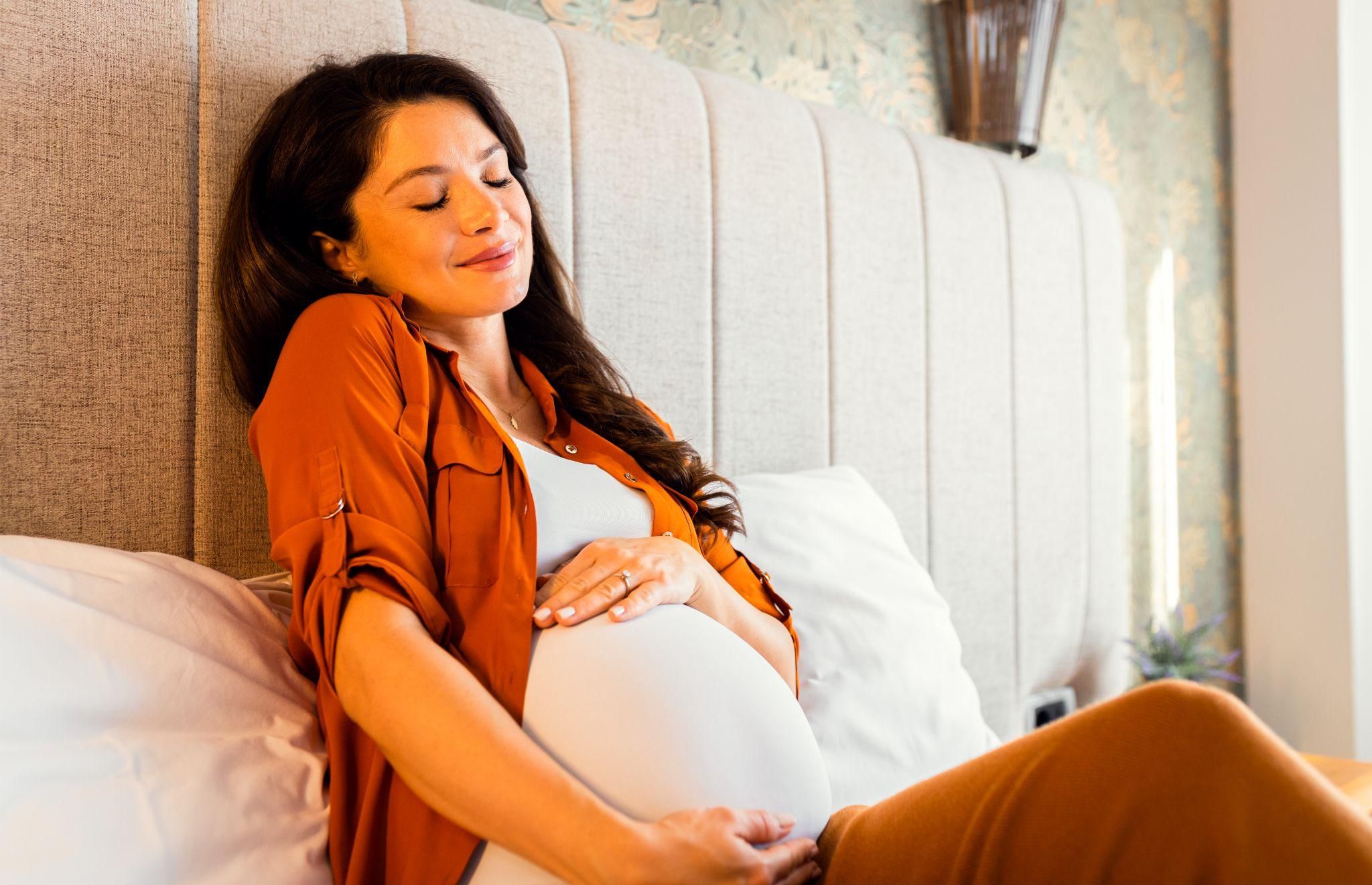 A pregnant woman relaxes on her bed holding her bump in her hands.