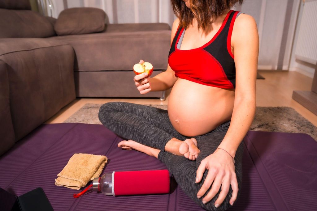 A pregnant woman chills out and eats an apple after doing antenatal yoga poses on a yoga mat.